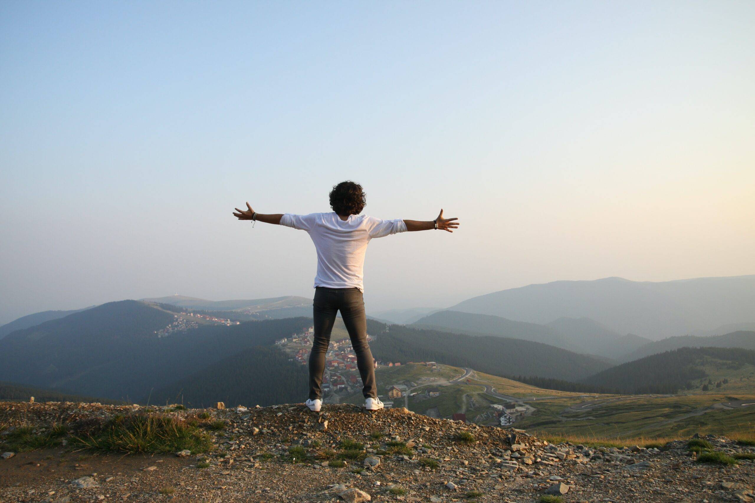 man standing on mountain top overlooking other mountains with his arms spread wide apart